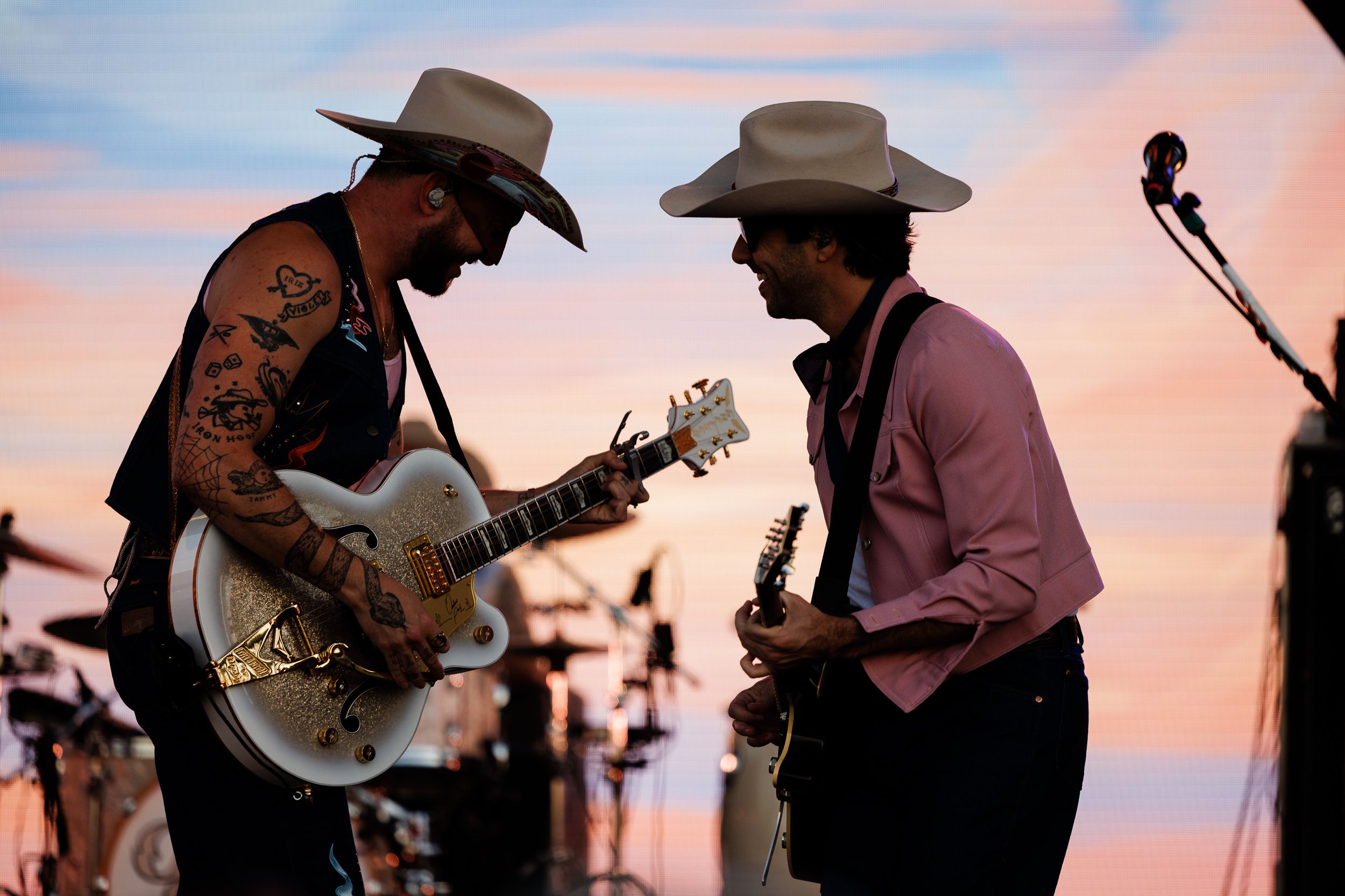 Two guitarists in cowboy hats facing each other against a pastel prairie sunset