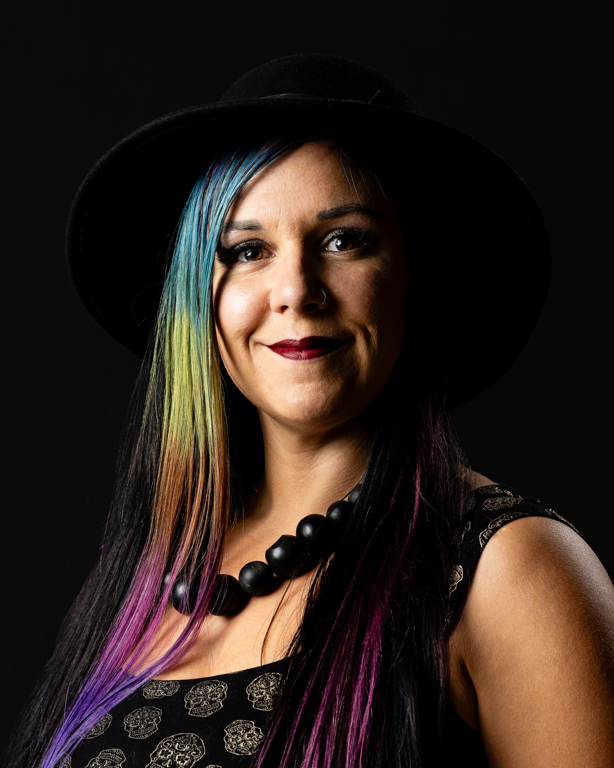 Dark-background portrait of a woman in a black hat with rainbow hair
