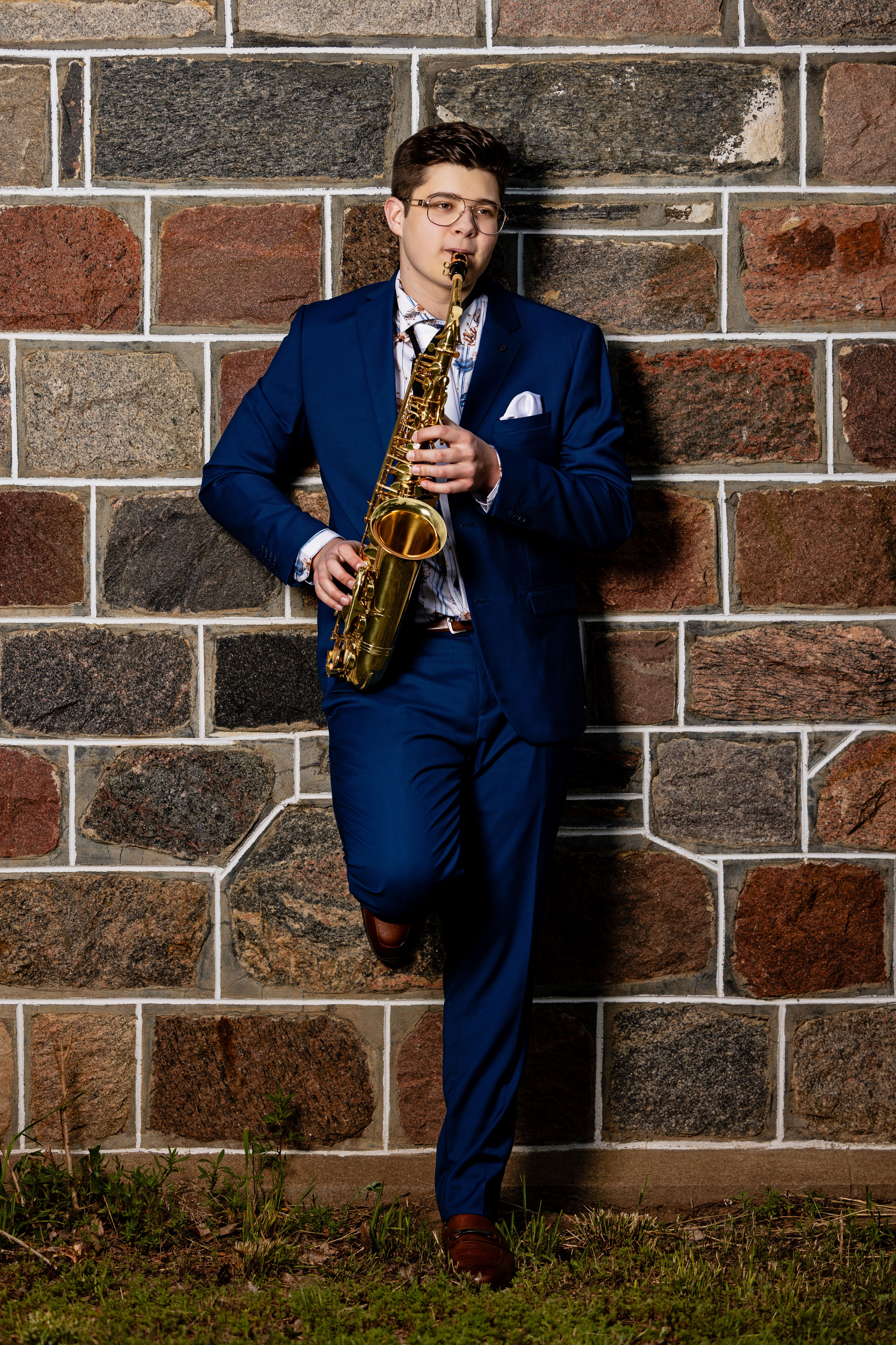 Grad in a blue suit with a saxophone against a fieldstone wall
