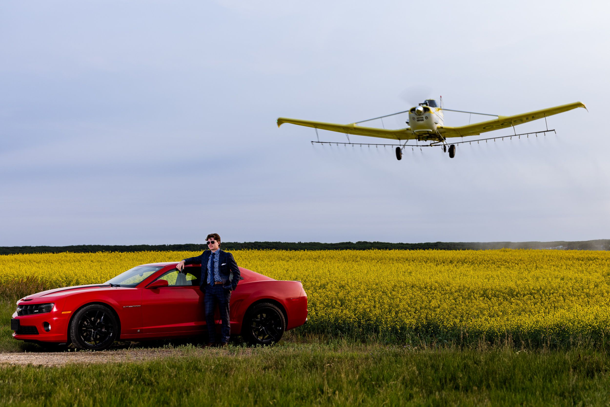Grad leaning on a red Camaro in a canola field while a crop duster flies overhead