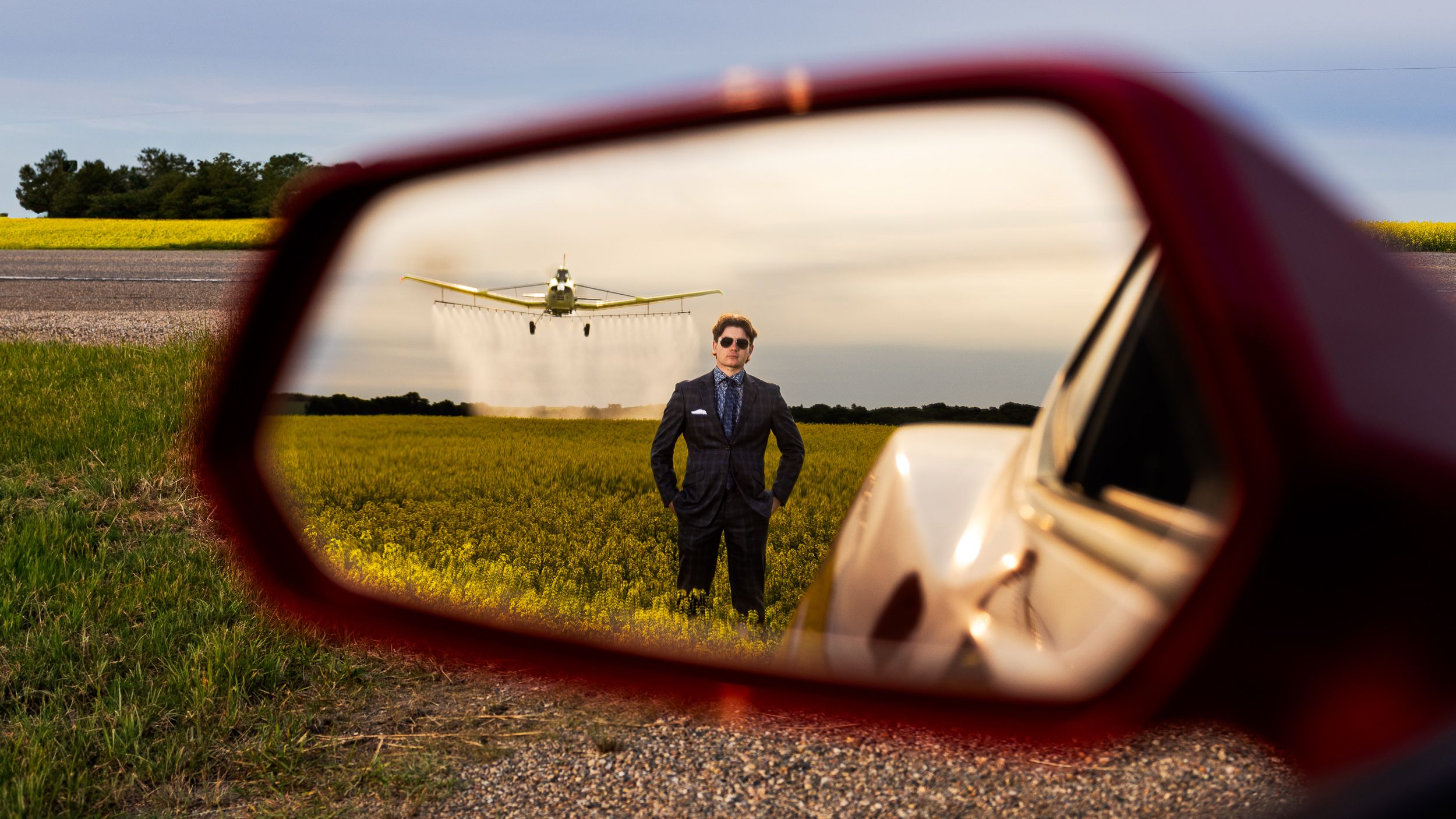 Grad in a blue suit reflected in a red Camaro side mirror in a canola field near North Battleford