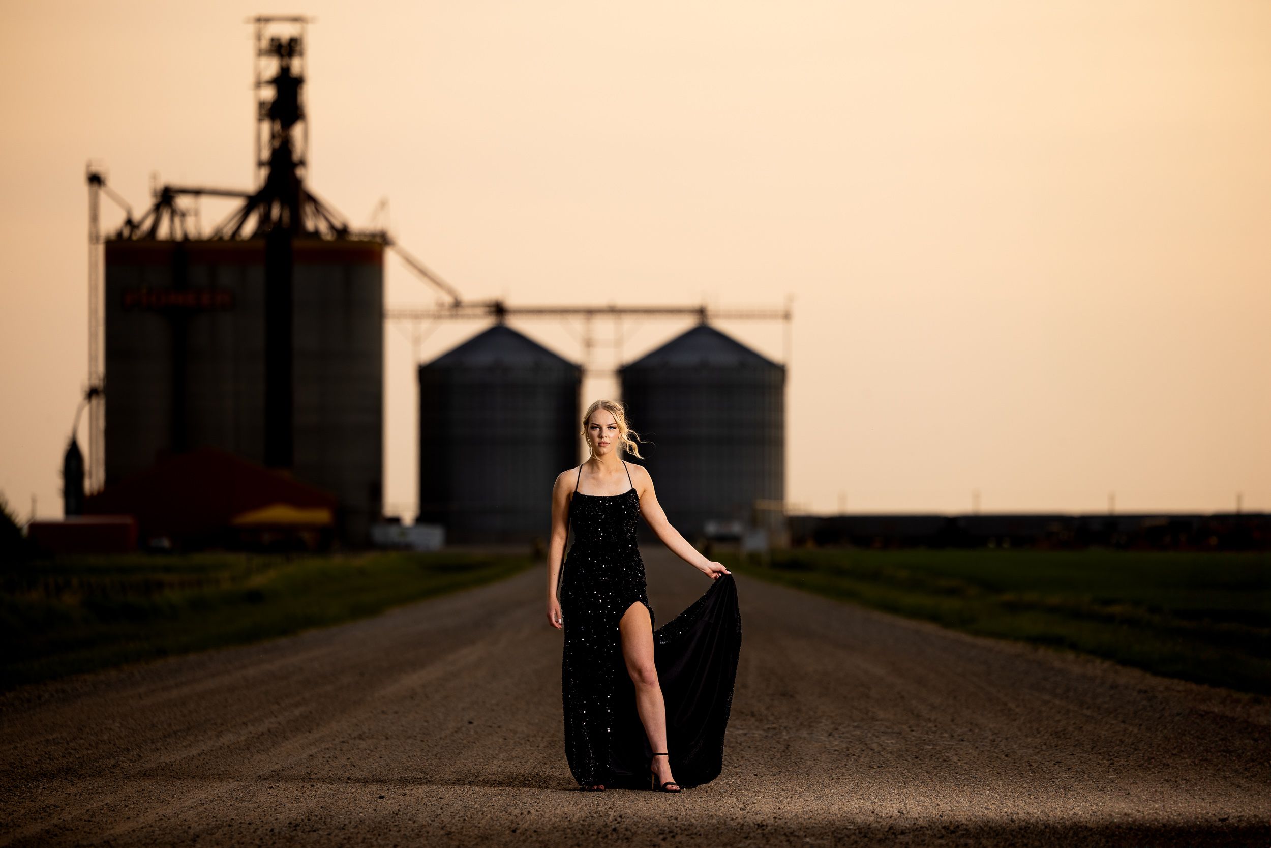 Grad in a black dress on a gravel road beside a prairie grain elevator at golden hour near North Battleford, Saskatchewan